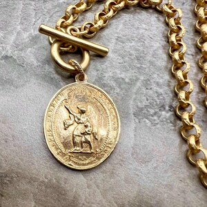 Pu&ograve; includere: Collana color oro con un grande ciondolo ovale con una figura di angelo. Il ciondolo &egrave; sospeso da una catena spessa con una chiusura a levetta. La collana &egrave; di un caldo colore oro.