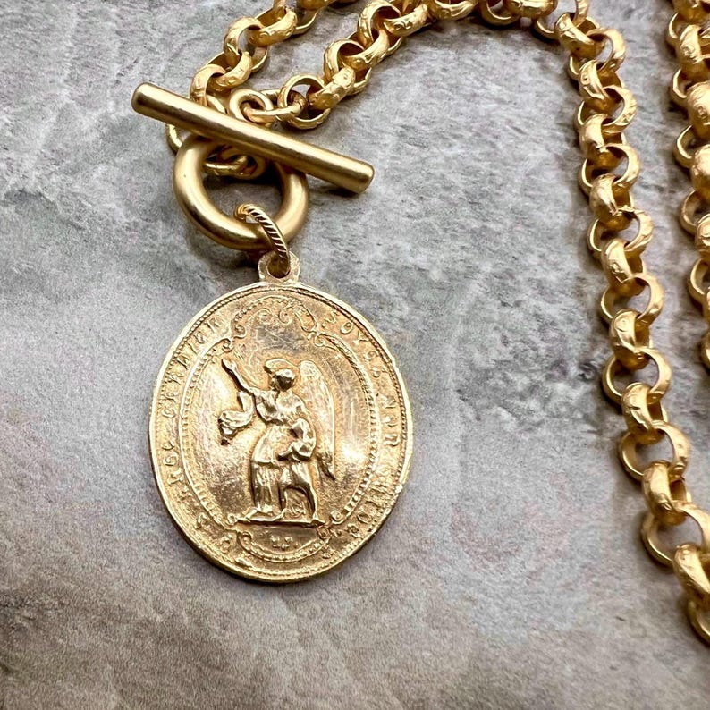 Pu&ograve; includere: Collana color oro con un grande ciondolo ovale con una figura di angelo. Il ciondolo &egrave; sospeso da una catena spessa con una chiusura a levetta. La collana &egrave; di un caldo colore oro.