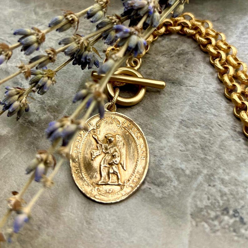 Pu&ograve; includere: Una collana color oro con un ciondolo rotondo raffigurante un angelo. La collana ha una chiusura a toggle e una catena spessa. Rami di lavanda sono disposti vicino alla collana su una superficie grigia. Il ciondolo ha un diametro di circa 2,5 cm.