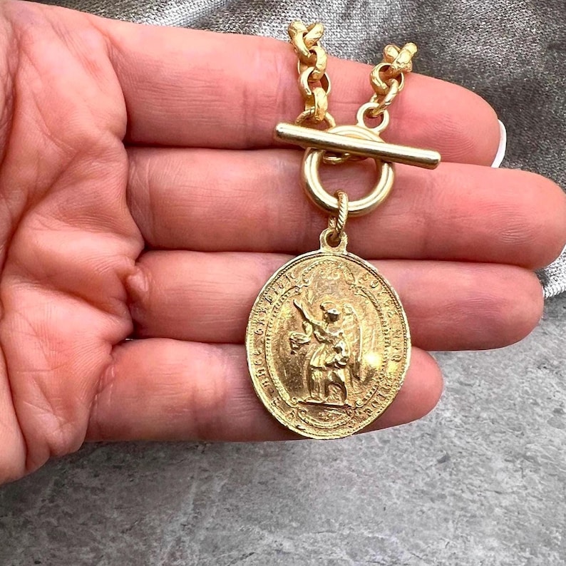 Pu&ograve; includere: Collana color oro con un grande ciondolo ovale con una figura incisa. Il ciondolo pende da una catena spessa con una chiusura a levetta. La collana &egrave; di un caldo color oro antico.