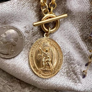 Pu&ograve; includere: Una collana color oro con un pendente rotondo raffigurante una figura di angelo. Il pendente &egrave; attaccato a una catena d'oro con una chiusura a levetta. Una moneta d'argento &egrave; accanto alla collana su un tessuto grigio.