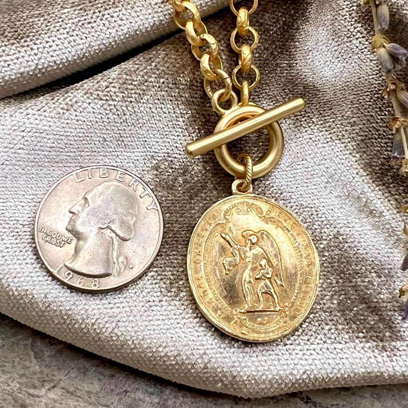 Pu&ograve; includere: Una collana color oro con un pendente rotondo raffigurante un angelo. La collana ha una chiusura a toggle e una catena spessa. Una moneta da 25 centesimi americana &egrave; accanto al pendente per il confronto delle dimensioni. La moneta &egrave; datata 1968.