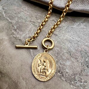 Pu&ograve; includere: Collana color oro con un pendente rotondo raffigurante una figura in rilievo. La collana ha una chiusura a toggle e una catena con un design a maglie grosse. Il pendente misura circa 3,8 cm di diametro.