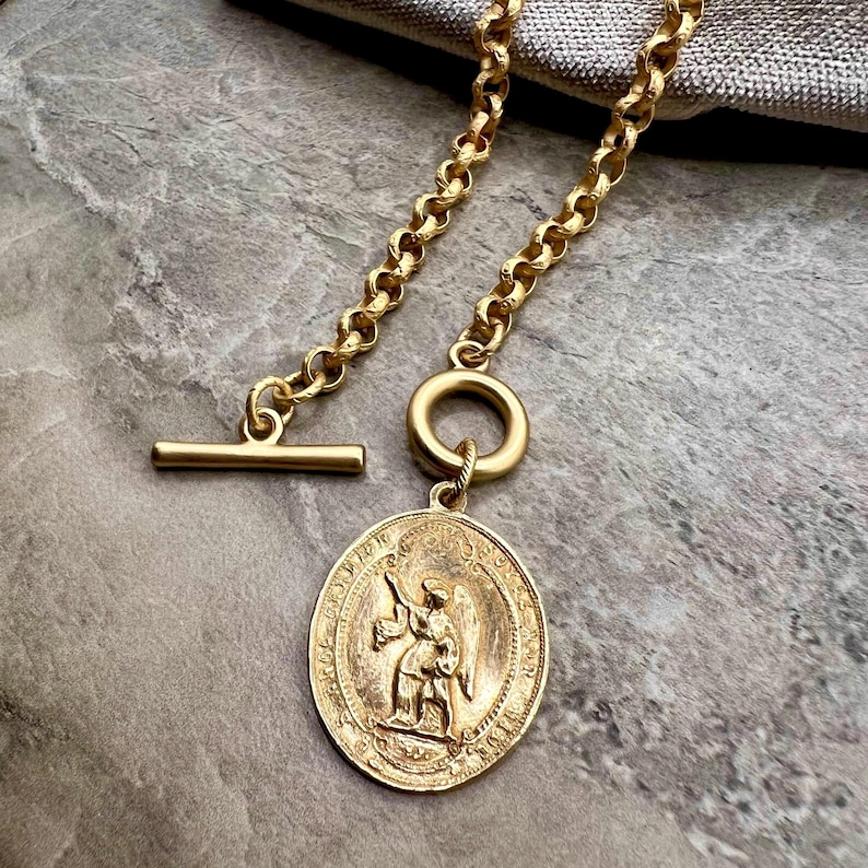 Pu&ograve; includere: Collana color oro con un pendente rotondo raffigurante una figura in rilievo. La collana ha una chiusura a toggle e una catena con un design a maglie grosse. Il pendente misura circa 3,8 cm di diametro.