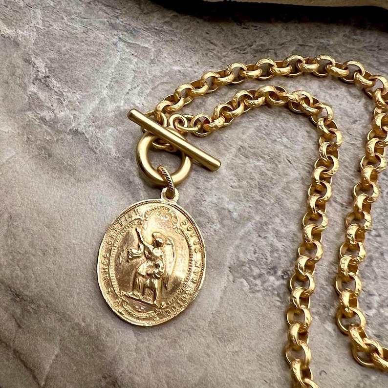 Pu&ograve; includere: Collana color oro con un pendente dettagliato raffigurante una figura e una chiusura a levetta. La catena ha un design a maglie rotonde e spesse. Il pendente &egrave; di forma ovale con dettagli intricati. La collana &egrave; esposta su una superficie strutturata e neutra.