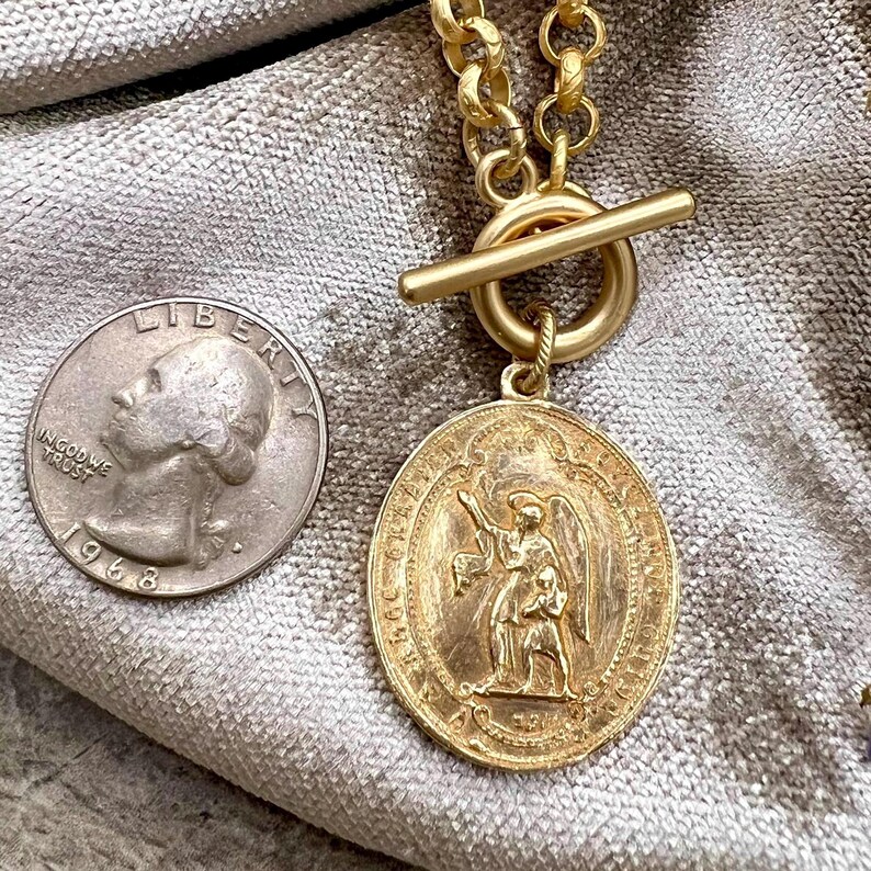 Pu&ograve; includere: Una collana color oro con un grande ciondolo ovale con una figura alata incisa. La collana ha una chiusura a levetta e una catena spessa. Una moneta &egrave; accanto al ciondolo per il confronto delle dimensioni.