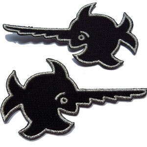 Puede incluir: Dos parches bordados negros para planchar con la forma de un pez espada estilizado. Los parches tienen un contorno plateado.