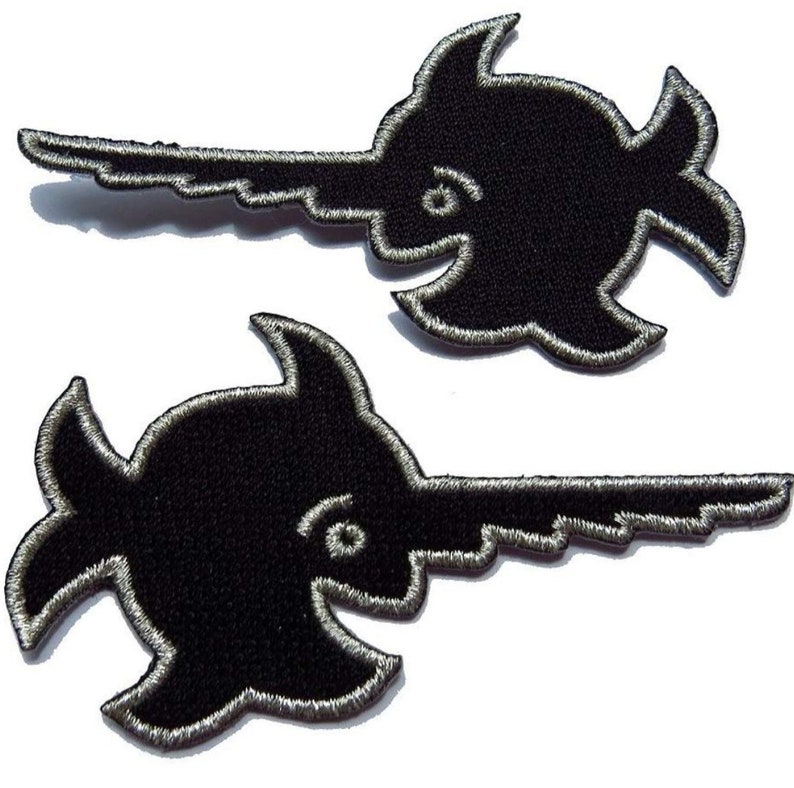 Puede incluir: Dos parches bordados negros para planchar con la forma de un pez espada estilizado. Los parches tienen un contorno plateado.