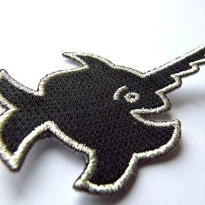 Puede incluir: Parche de hierro negro bordado de un pez espada estilizado con contorno plateado.