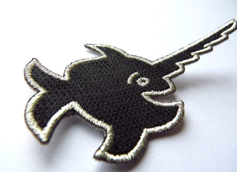 Puede incluir: Parche de hierro negro bordado de un pez espada estilizado con contorno plateado.