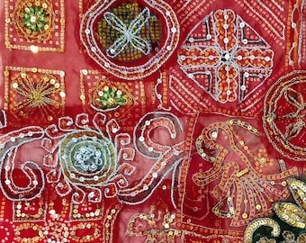 May include: Close-up of a red textile with intricate embroidery and sequin embellishments. The design features geometric shapes, floral patterns, and a figure, all in gold, silver, and green. The fabric has a sheer quality.