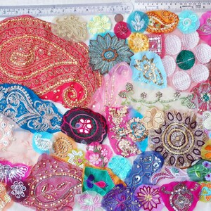 May include: A collection of colorful fabric embellishments, including paisley shapes, flowers, and abstract designs. The pieces are adorned with sequins, beads, and embroidery in shades of red, blue, pink, and gold. A ruler is visible along the top and left edges.