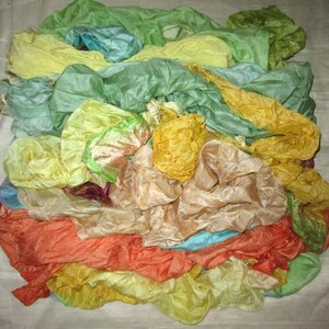 May include: A pile of colourful silk scarves in shades of green, yellow, orange, and blue. The scarves are arranged in a loose, flowing heap, creating a sense of movement and texture.