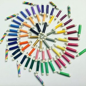 May include: A collection of colorful pencil-shaped charms with white tips and silver jump rings. The charms are arranged in a circle on a white background.