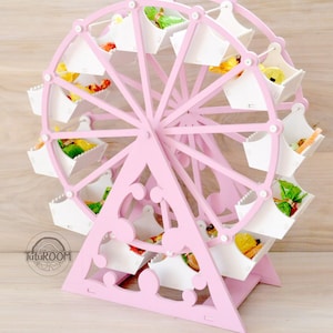 Puede incluir: Porta golosinas de madera de color rosa en forma de noria con compartimentos blancos. La noria tiene 12 compartimentos y está decorada con puntos blancos. La noria está sobre una base de madera.