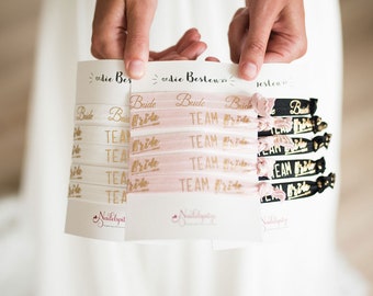Op de afbeelding: Een set roze, witte en zwarte elastiekjes voor haar met gouden letters. De elastiekjes hebben de woorden "Bride" en "Team Bride" erop gedrukt.