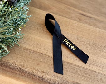 Könnte beinhalten: Ein schwarzes Band mit dem Namen "Peter" in goldenen Buchstaben. Das Band ist zu einer Schleife gebunden und liegt auf einer Holzoberfläche.