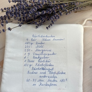 Può includere: Una ricetta scritta a mano per Eierlikörkuchen (torta al liquore tedesco) su uno sfondo di tessuto bianco. La ricetta include ingredienti come uova, zucchero, farina, margarina, zucchero vanigliato, lievito in polvere, liquore e codette di cioccolato. La ricetta indica di cuocere la torta in uno stampo da plumcake a 180 gradi Celsius per 60-70 minuti.