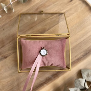 May include: A glass box with a gold frame and a pink velvet pillow inside. The pillow has a small, round, gold-colored charm with a white design on it. A pink ribbon is attached to the charm.