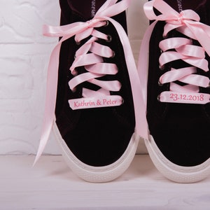 Könnte beinhalten: Ein Paar dunkelburgunderfarbene Samt-Sneaker mit rosa Satinbändern, die zu einer Schleife gebunden sind. Der linke Schuh hat ein Etikett mit der Aufschrift "Kathrin & Peter" und der rechte Schuh hat ein Etikett mit der Aufschrift "23.12.2018".