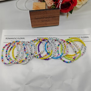 Puede incluir: Una colección de pulseras de cuentas de colores con letras blancas que deletrean nombres y palabras como "VOTE" y "HARRIS 2024". Las pulseras están sobre una superficie blanca, con una caja de madera y un arreglo floral en el fondo.