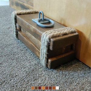Può includere: Un fermaporta in legno con una maniglia in corda e un anello in metallo. Il fermaporta è marrone e ha un aspetto rustico.