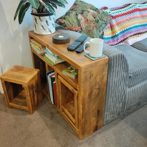 May include: A rustic wooden side table with two shelves and a small stool. The table is attached to a grey sofa and has a white mug and remote controls on top.