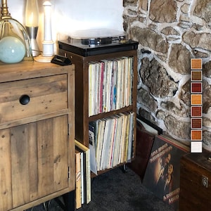 May include: A wooden record player stand with two shelves for storing vinyl records. The stand is dark brown and has a black turntable on top. The stand is next to a wooden cabinet with a drawer.