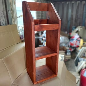 May include: A tall, wooden shelf unit with a reddish-brown stain. The unit has three shelves and a top section with a handle-like design. The shelves are spaced apart, and the unit is made of solid wood.