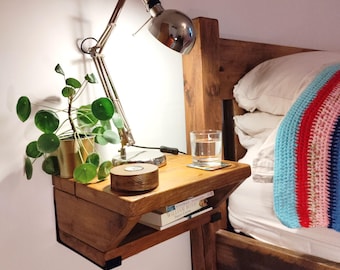 May include: A wooden bedside table with a shelf, attached to the wall next to a bed. A lamp with a silver shade is on the table, along with a glass of water and a book titled "Eleanor Oliphant is Completely Fine" by Gail Honeyman.