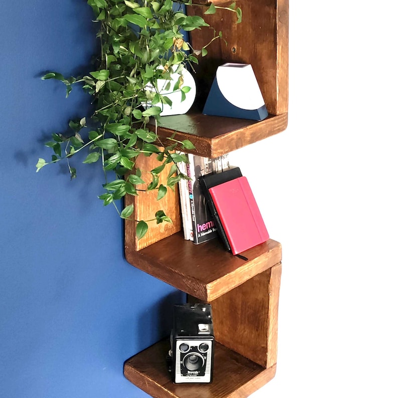 May include: A wooden wall shelf with a staggered design. The shelf is stained a dark brown color and has three shelves. The top shelf has a white and blue geometric vase, the middle shelf has a stack of books and a red journal, and the bottom shelf has a vintage camera. A green plant is hanging from the top shelf.