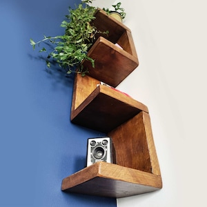 May include: A set of wooden shelves in a geometric shape, mounted on a blue wall. The shelves are made of dark wood and have a natural finish. The shelves are empty, but there is a small camera on the bottom shelf.