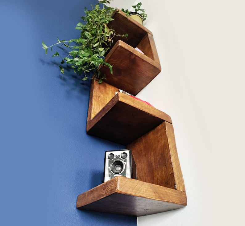 May include: A set of wooden shelves in a geometric shape, mounted on a blue wall. The shelves are made of dark wood and have a natural finish. The shelves are empty, but there is a small camera on the bottom shelf.