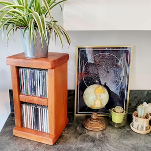 May include: A wooden CD storage cabinet in a warm, reddish-brown finish, holding multiple CDs. A potted plant sits on top. A framed star chart, a globe, and small potted cacti are also visible.