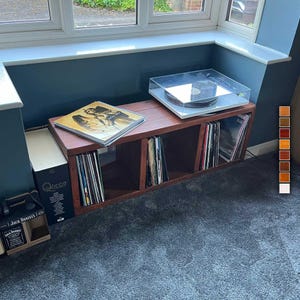 May include: A wooden record shelf with four compartments, each filled with vinyl records. A turntable with a clear dust cover sits on top of the shelf. The shelf is in front of a window with a blue wall behind it.