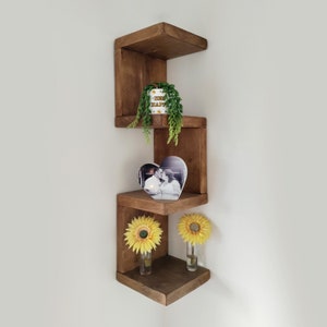 May include: A set of three wooden corner shelves in a dark brown stain. The shelves are stacked in a staggered pattern, creating a unique and modern look. The shelves are decorated with a small potted plant, a heart-shaped photo frame, and two yellow sunflowers in vases.