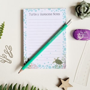 May include: A green pencil with an eraser on the end is lying on top of a notepad with the text "Turtle-y Awesome Notes" printed on the front. The notepad has a blue and white background with a turtle and coral reef design. There is a purple eraser with the text "we all make mistakes" printed on it.