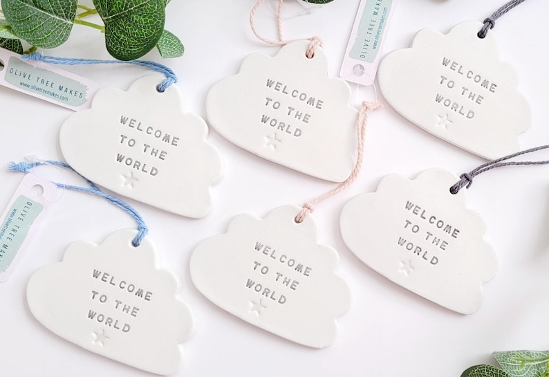 May include: Cloud-shaped ceramic ornaments in white, each bearing the text "WELCOME TO THE WORLD" in grey. They feature a star detail and are strung with coloured cords. Ideal as a gift for a new baby or baby shower.
