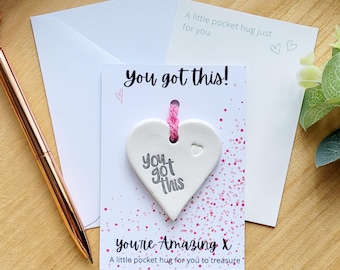 May include: A white heart-shaped pocket hug with the text "You got this!" and "you're Amazing X" on a card. The card includes a pink string and a small heart. A white envelope and a pen are also in the image.