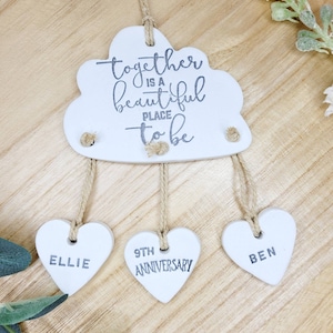 May include: White ceramic cloud with the text "together is a beautiful place to be" hanging from twine with three white ceramic hearts that say "Ellie", "9th Anniversary", and "Ben".