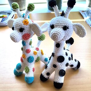 Puede incluir: Dos juguetes de jirafa de crochet. Una jirafa es blanca con lunares negros y botones negros. La otra jirafa es blanca con lunares de colores. Ambas jirafas tienen orejas y narices negras.