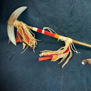 Può includere: Un'antica arma cerimoniale dei nativi americani con una lama curva, un'asta di legno decorata e ornamenti di piume e pelliccia. La lama è di colore grigio chiaro e l'asta è avvolta con spago e dipinta con strisce rosse, gialle e bianche.