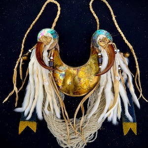 May include: A handcrafted necklace featuring a brass crescent, adorned with large claws, iridescent accents, and white fur. The necklace includes beaded strands and leather ties, set against a black background. The design incorporates natural elements.