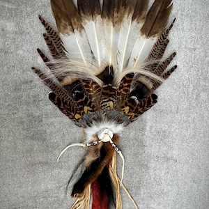 Peut inclure: Éventail artisanal composé de plumes brunes, blanches et noires. Le motif central de l'éventail comprend des plumes à motifs, de la fourrure, des perles et des liens en cuir. La poignée est enveloppée de tissu rouge et marron. Environ 25 cm de long.
