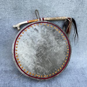 May include: A circular, grey drum with a red and orange dotted border. A drumstick with a bone handle and black and brown tassels rests on the drum. A traditional musical instrument.