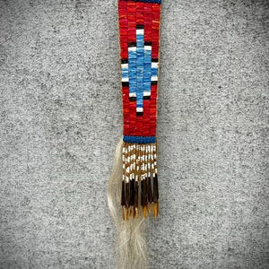 May include: A rectangular, beaded pendant with a red background and a central diamond pattern in blue, white, and black. The top edge is blue, and the bottom features a fringe of white, brown, and tan strands, with a long, light-colored hair tail.
