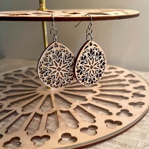 Könnte beinhalten: Paar Holz-Ohrringe in Tropfenform mit filigranen floralen Ausschnitten. Die Ohrringe sind hellbraun und haben silberfarbene Ohrhaken. Sie werden auf einem dekorativen Holzständer mit floralem Design präsentiert.