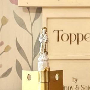 May include: A small, white statue of a religious figure holding a child, with gold accents. The statue is mounted on a gold hinge, with the word "Toppe" visible in the background.