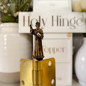 Könnte beinhalten: Ein goldenes Türscharnier mit einer kleinen, detaillierten Bronzefigur eines Menschen, der eine Robe trägt und ein Buch hält. Die Figur steht auf dem Stift des Scharniers. Das Scharnier ist mit "Holy Hinge" und "Topper" beschriftet.