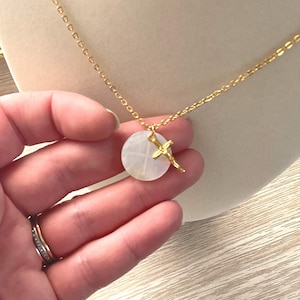 Op de afbeelding: Een gouden ketting met een ronde witte hanger met een gouden kruis. De hanger heeft een delicaat kruisontwerp.